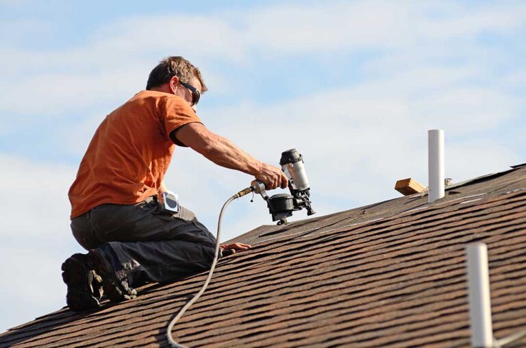 Person inspecting roof shingles and gutters from the ground after a storm, with branches and debris nearby.